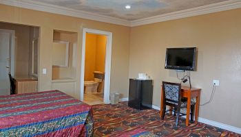 A hotel room with two beds, a TV on the wall, a desk with a chair, a minifridge, and a bathroom entrance. The room has carpeted flooring.