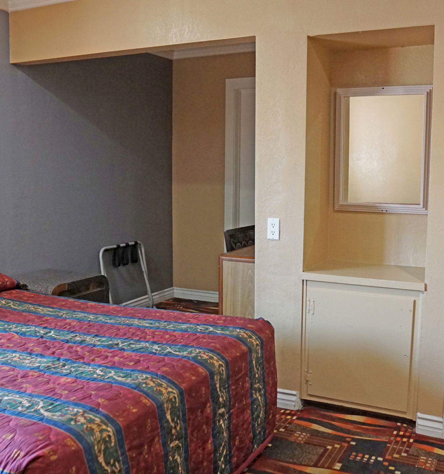 The image shows a hotel room with a bed, a small closet, a mirror, and a bathroom with towels hanging. The room has simple furnishings.