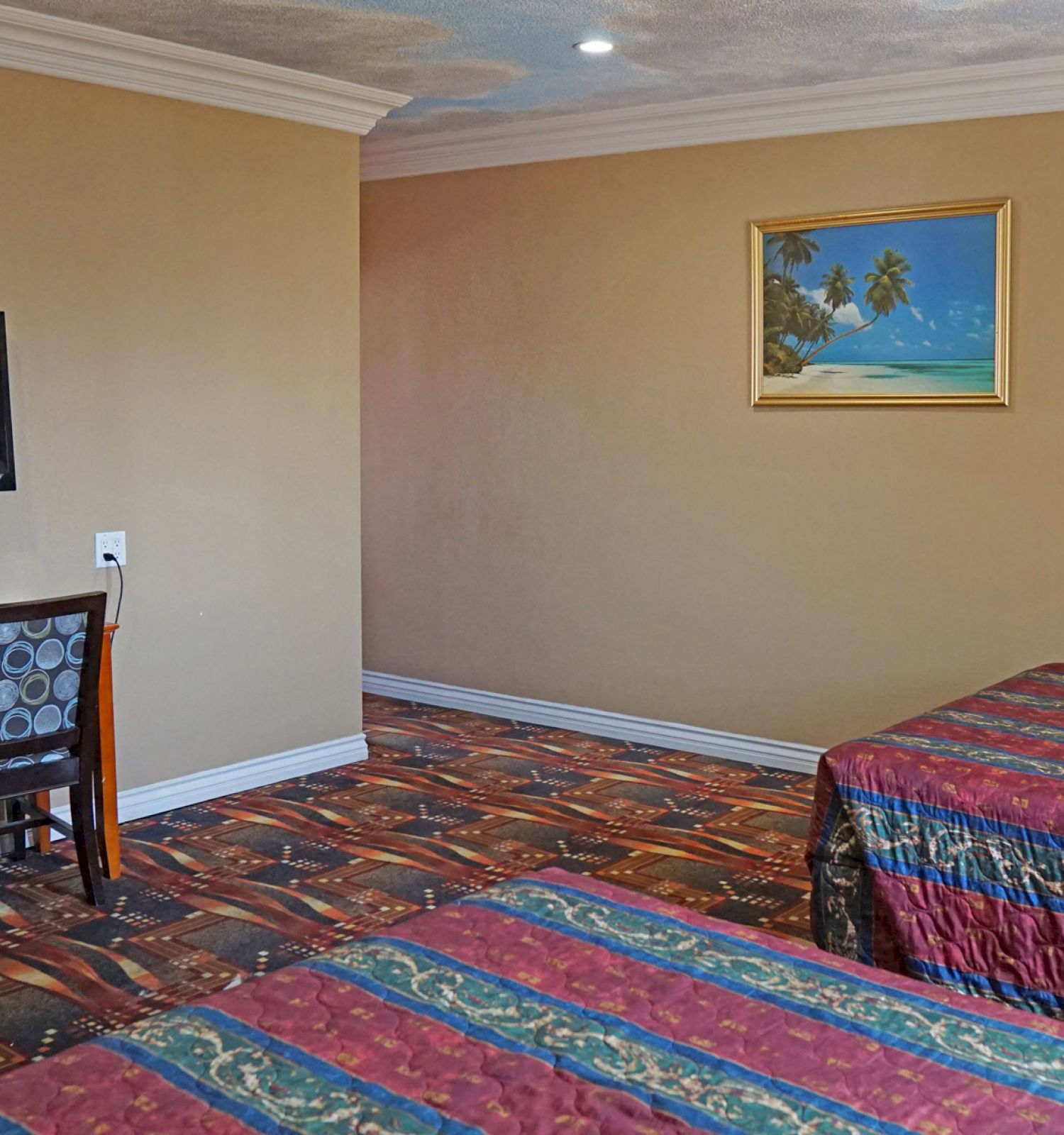 A hotel room with two beds, a desk with a chair, a wall-mounted TV, and a tropical beach painting on the wall.