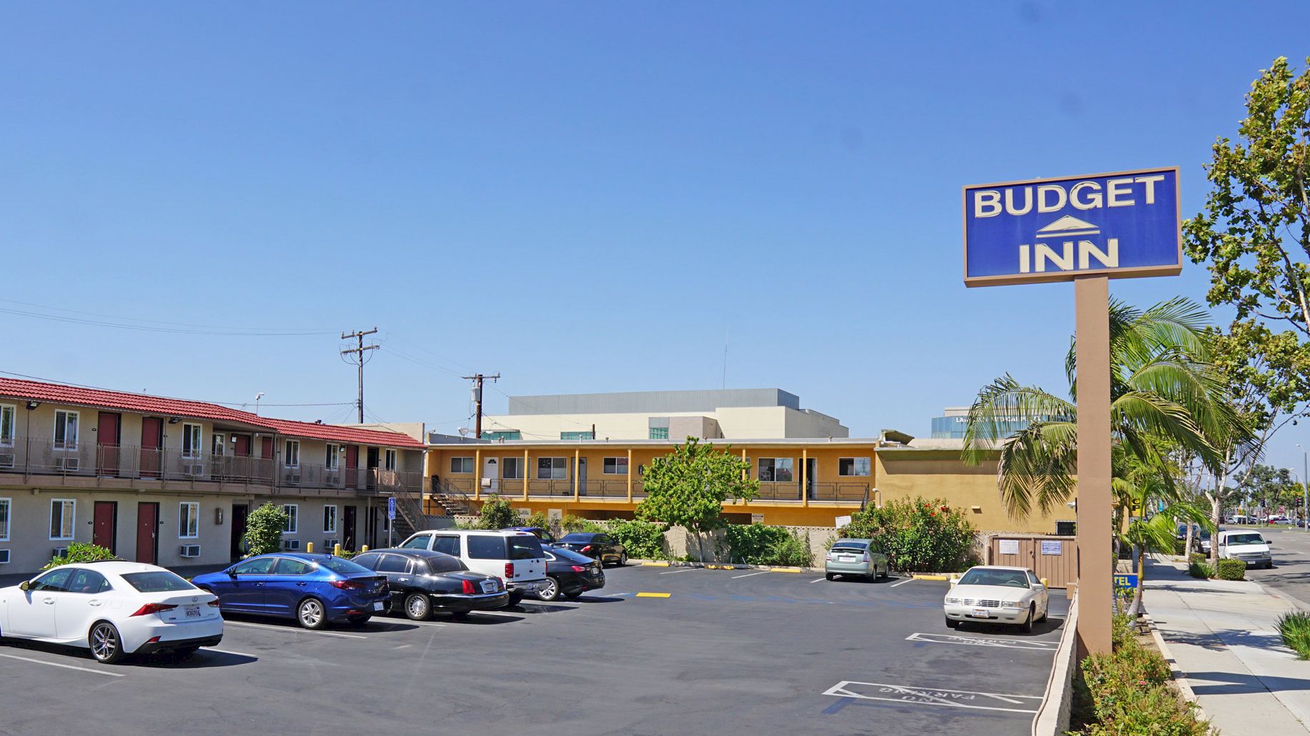The image shows a "Budget Inn" motel with a parking lot area and several cars parked in front of a two-story building on a clear day.